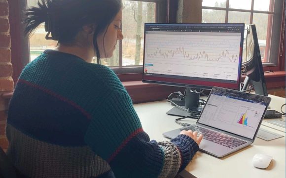 Woman seated at a desk working on a laptop with a large monitor and two windows.