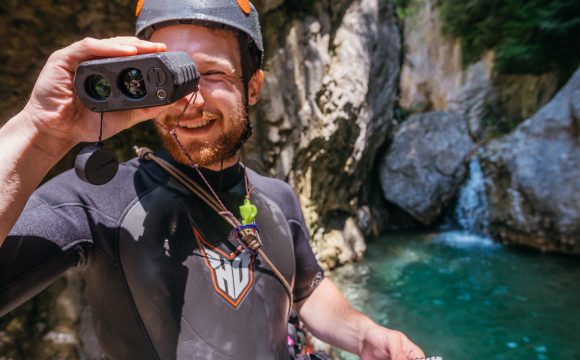 An image of a man wearing a wetsuit and helment standing infront of a small waterfall holding a scientific instrument up to his eyes