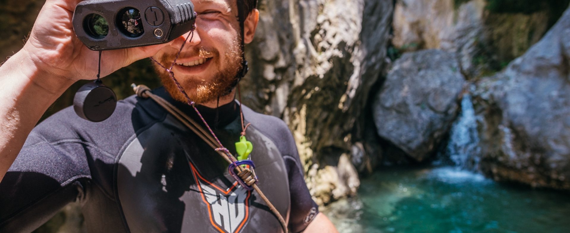 A man wearing a wet suit and helmet in front of a body of water holding a binocular like instrument up to his face